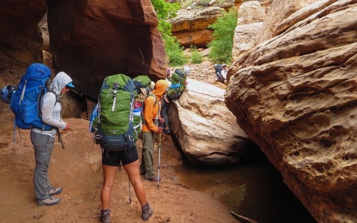 A group of people wearing backpacks hike through a canyon among large boulders.